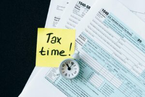 Une pile de formulaires fiscaux avec une horloge et un post-it jaune indiquant « C'est l'heure des impôts ! » indiquant l'urgence.